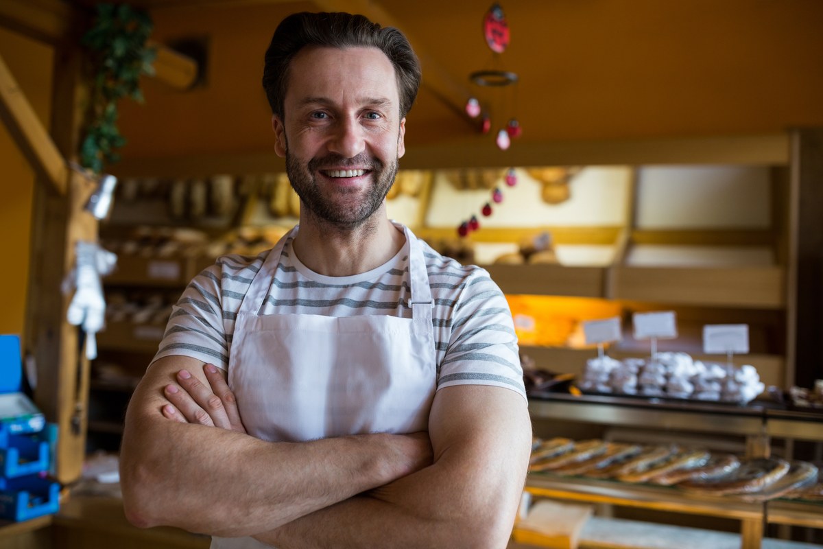 Een trotse bakkerijeigenaar die met gekruiste armen glimlacht in zijn eigen winkel